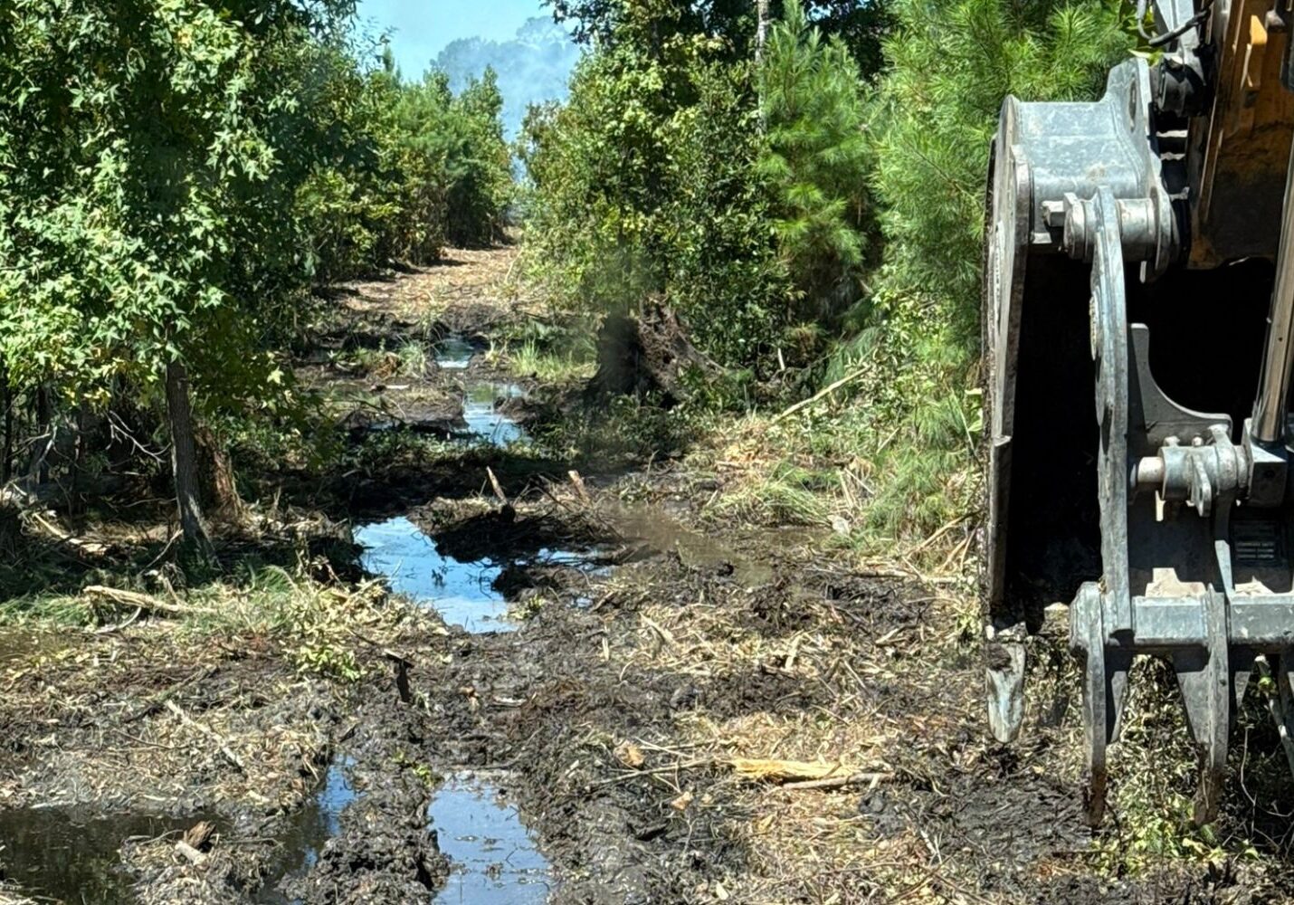Muddy cleared pathway through a wooded area with standing water and disturbed earth, flanked by green trees, with the arm and bucket of a construction excavator visible on the right side.